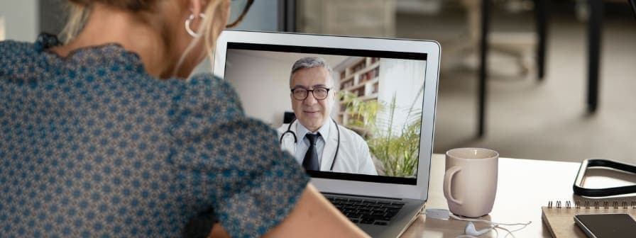 Young woman sits facing toward a laptop screen, talking with doctor in a white coat on the screen, representing a virtual health visit